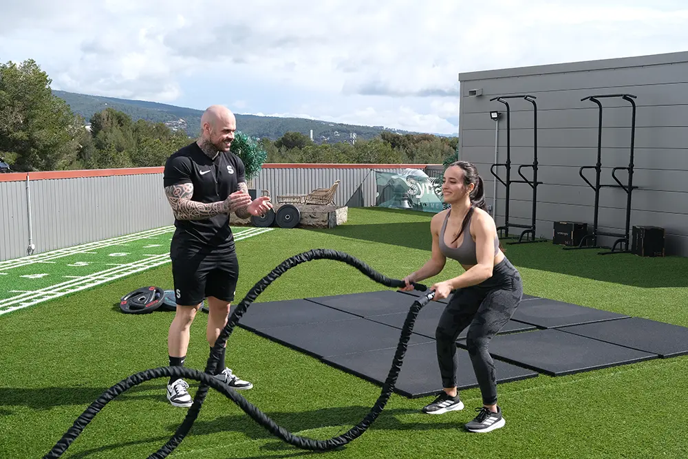 Fitness trainer motivating a female client during a battle ropes workout, utilizing body composition analysis to track progress beyond weight.